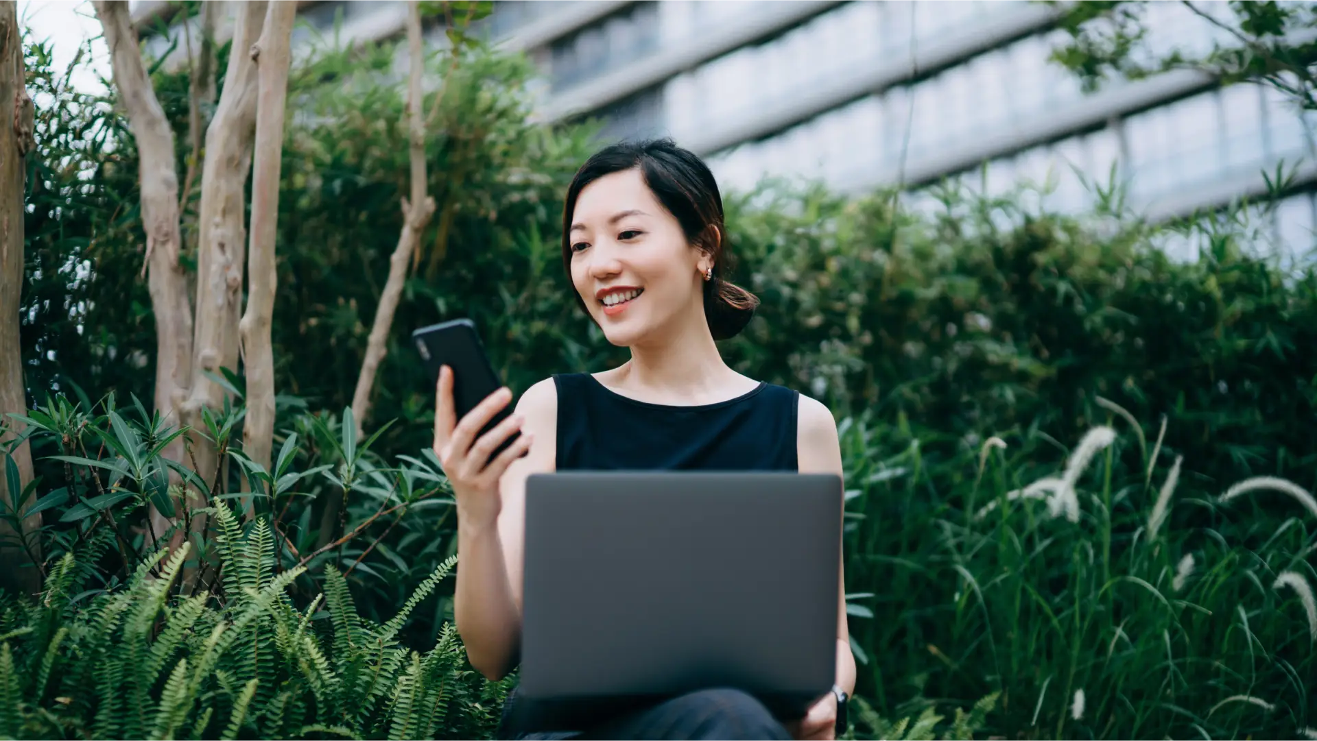 A woman looking at her phone and holding a laptop in a garden